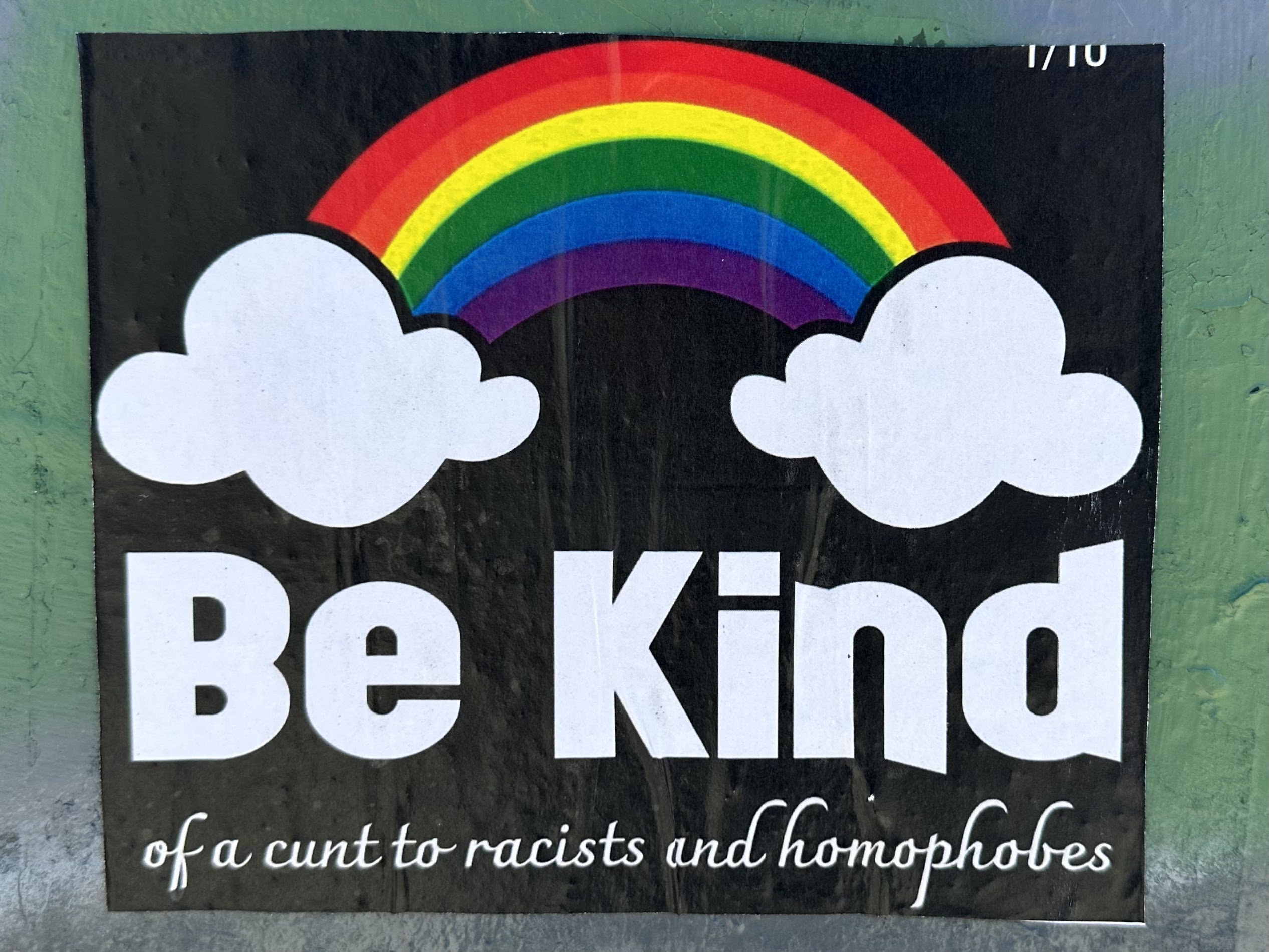 Wall sticker with words under a rainbow connecting two fluffy white clouds. Written in large letters is, “Be Kind”. Underneath them in a cursive script it continues, “of a cunt to racists and homophobes”.