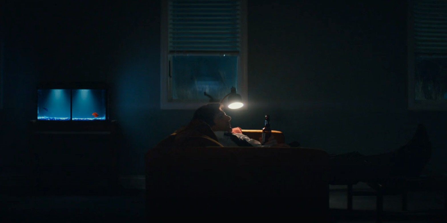 Screenshot from TV series Severance. The main character Mark sits on his couch with a single lamp turned on next to him. He is slumped back, his feet up on the table in front of him and holding a beer. Behind him there is a fish tank with a blue and a red fish inside.